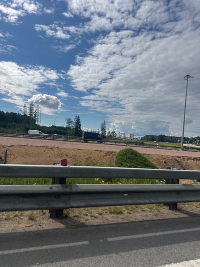 The image depicts a roadside scene in Levashovo, Russia, under a partially cloudy sky. The perspective is from the side of a road, with a metal guardrail in the foreground dominating the right side of the frame. The guardrail consists of multiple horizontal metal bars, casting shadows on the adjacent asphalt road surface.

Beyond the guardrail, a dirt path runs parallel to the road, lined with sparse vegetation and some small white flowers. In the mid-ground, a dark-colored van is visible on what appears to be a construction or unpaved road. Further into the background, a line of coniferous trees forms a dense forest. Tall poles with lights, typical of road lighting, are also present in the distance. The sky is a vibrant blue, interspersed with wispy white clouds that are partially obscuring a brighter sky. One large, cumulus cloud formation is particularly prominent.

The scene suggests a daytime setting, likely late morning or early afternoon, given the bright light and shadows. The weather appears to be pleasant with a mix of sun and clouds. There are no people visible in the image. The overall impression is one of an industrial or developing area adjacent to a natural landscape. No text is visible in the image.