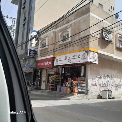 The image depicts a street scene, apparently in the city of Nuseirat in the Palestinian Territories, as implied by the signs in Arabic, which include the words "فلسطين" and "غزة". The view is from inside a vehicle, showing the lower edge of a car door and window. The focus is on a storefront labeled "فرج الله ماركت", which appears to be a small supermarket. Above this store is another sign, also in Arabic, with the same name. Beneath the supermarket is a "coredoo" sign, which might be a brand or service. Adjacent to the market, other businesses are discernible, including what seems to be a mobile phone shop ("فاصيلنة الجوال"), and further down, a pharmacy ("صيدلية د. صافي"). The building facade shows signs of aging with some graffiti. A complex network of electrical wires is visible overhead. The surrounding buildings are old, with traditional architectural elements. The lighting suggests it is daytime. The overall impression is of a typical street scene in a densely populated urban area.