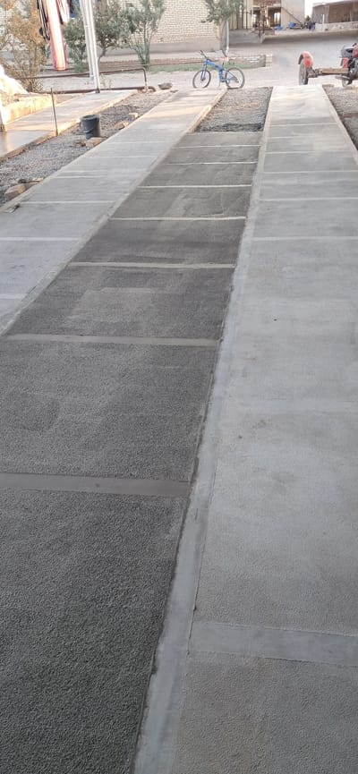 The image shows a ground-level view of a paved area, possibly a walkway or parking lot, in Mary, Turkmenistan. The paving is composed of large, rectangular concrete slabs with visible seams between them. Some of the slabs have a rough, pebbled texture, while others appear smoother. A narrow strip of gravel runs alongside the paved area.
In the background, on the left side of the image, a blue bicycle is leaning against something. Further back, a motorcycle is parked. There are also glimpses of buildings and trees, suggesting an outdoor setting with some signs of development or construction. The lighting indicates it is either early morning or late afternoon. The weather appears to be clear.
The scene appears to be a quiet, perhaps residential or communal area. There are no people clearly visible in the frame. The overall impression is one of a functional, outdoor space. No text is visible in the image.