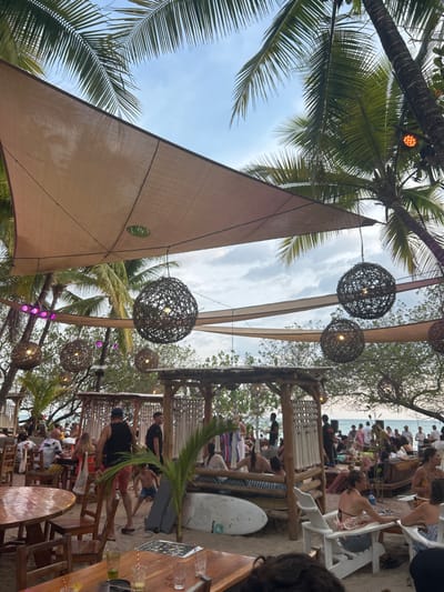 The image captures a scene at a beachfront restaurant in Santa Teresa, Costa Rica. The location, determined by the provided coordinates (latitude: 9.632, longitude: -85.158), is a popular tourist destination known for its beautiful beaches and relaxed atmosphere.
A large canopy, stretched over wooden posts, provides shade for a bustling crowd. The canopy is adorned with woven, spherical lamps hanging from its corners. The restaurant is situated on a sandy surface, with a lush, tropical backdrop of palm trees. Patrons are seated at various tables and chairs throughout the area, enjoying drinks and food.
Some people are engaged in conversation, while others are gazing out at the ocean. There is a surfboard resting on the sand, near a wooden lounge chair. The scene is characterized by a vibrant and casual ambiance, consistent with the laid-back atmosphere of Santa Teresa.