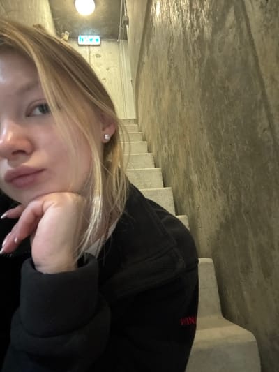 An indoor scene in Rosh Haayin, Israel, features a partial close-up of a young adult female on the left, with an ascending concrete stairwell in the background. The female's left profile, including her eye, nose, and slightly pouted mouth, is visible. Her blonde hair frames her face. She wears a small, light-colored stud earring in her left earlobe and a thin, copper-toned necklace over a white inner shirt. Her left hand, adorned with a French manicure, rests with her fingers under her chin. She is dressed in a black, possibly fleece, long-sleeved top, which displays red embroidered or printed text, possibly "WIN," on the lower right front. The background consists of a concrete staircase with at least six visible steps ascending towards the upper left. The surrounding walls and ceiling are also exposed, rough concrete. An overhead circular light fixture and a green emergency exit sign with white pictograms are visible on the ceiling and upper left wall, respectively. Exposed conduits run along the ceiling.