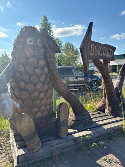 The image depicts a whimsical, large wooden sculpture of a pinecone character sitting outdoors in the city of Vyra, Russia. The sculpture has a smiling face with circular eyes and an owl perched on its head. To the right of the pinecone character, a wooden sign shaped like an arrow points to the left and reads "ВЫСТАВКА," which translates to "EXHIBITION." The sculpture is seated on a wooden platform made of crates and planks. In the background, a dark-colored SUV is visible parked behind a chain-link fence, with trees and a building also in the distance. The scene is illuminated by bright sunlight under a blue sky with scattered clouds.
