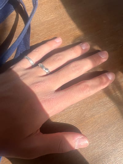 The image displays a human left hand, palm down, resting on a light-colored wooden surface. The hand exhibits light-toned skin with visible texture. Two silver-colored rings adorn the fourth digit: a thin, twisted band is positioned distally on the proximal phalanx, while a thicker, irregularly contoured band is located proximally to it on the same phalanx. The fingernails are unpolished, showing natural appearance, visible cuticles, and minor surface irregularities. Direct sunlight illuminates the scene, casting distinct shadows from the hand onto the wooden surface. A dark blue fabric strap is partially visible in the upper left quadrant of the image. The location is Hof Ashkelon Regional Council, Israel.