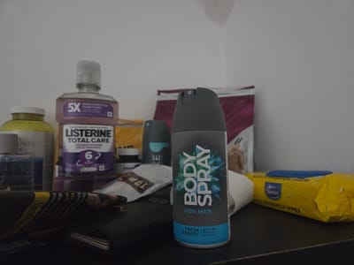The scene is located in the city of Braine-l'Alleud, Belgium. It depicts an indoor arrangement of personal care and household items positioned on a dark, flat surface against a plain, light-colored wall. In the central foreground, a grey aerosol can of "BODY SPRAY FOR MEN" with "FRESH ENERGY" branding and a blue/green design is prominently displayed. To the left and slightly behind the body spray, a purple bottle of "LISTERINE TOTAL CARE" mouthwash is visible, featuring "5X MORE EFFECTIVE THAN FLOSS" and "6 IN 1 PROTECTION" text. Behind the Listerine bottle, a yellow container, likely a supplement bottle, is partially visible. Further left, a blue container is partially obscured. A dark-colored wallet with some papers or receipts on top rests in the left foreground. To the right of the Listerine, a dark grey/black deodorant container with a light blue accent is present. In the right mid-ground, a yellow package of "Spaargo" brand "HOUSEHOLD WIPES" is visible. Various soft packages and pouches are scattered in the mid-to-background, including a light-colored pouch with "TOP" visible text and a larger magenta-colored bag further back. No individuals are present, and no actions or interactions are occurring.