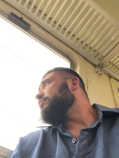 A man with a beard and dark hair is captured in a low-angle, close-up shot, seemingly taken from within a mode of transport like a train or bus, given the presence of a slatted overhead panel and a window. The man is looking down, his face partially obscured by his beard, with his eyes closed or looking downwards, suggesting a moment of rest or introspection. He is wearing a dark shirt with a repeating pattern that appears to be a logo, possibly a designer brand.
Visible text in the image includes Russian words. On a strip near the window, "АВАРИЙНЫЙ ВЫХОД" (Emergency Exit) is clearly legible, along with the word "ВЫХОД" (Exit) again on a red sticker. This indicates the location is likely a public transportation vehicle operating in a Russian-speaking region. The lighting suggests it might be daytime, with bright light coming from the window. The overall atmosphere is candid and personal.