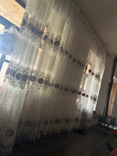 A close-up, eye-level shot captures a room with sheer, embroidered white curtains as the primary subject. The curtains, adorned with a recurring circular motif in dark blue and gold intricate patterns, are drawn partially across a window. Sunlight filters through the sheer fabric, creating a soft, diffused illumination. To the left, a plastic bag filled with items hangs near the window. In the background, to the right of the curtains, a dark metal wood-burning stove stands on a wooden platform. A wooden door is visible behind the stove. The floor appears to be dark wood. The scene is located in Mıxlıqovaq, Azerbaijan.