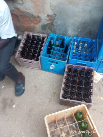 In Dokan Tofa, Nigeria, a low-angle, outdoor scene depicts a collection of bottle crates on a dusty ground. In the foreground, a tan plastic crate contains a few green glass bottles. To the right, a brown plastic crate is filled with empty dark brown glass bottles. Behind these, two blue plastic crates are visible. The left blue crate is filled with empty dark brown glass bottles, while the right blue crate holds a mix of full and empty dark brown and clear glass bottles, some with yellow labels. A person, partially visible on the far left, is wearing a white t-shirt and dark pants, with one foot shod in a black sandal. The background shows a textured, reddish-brown wall and a grey concrete structure. The image is dated "06/16/2026 15:55".