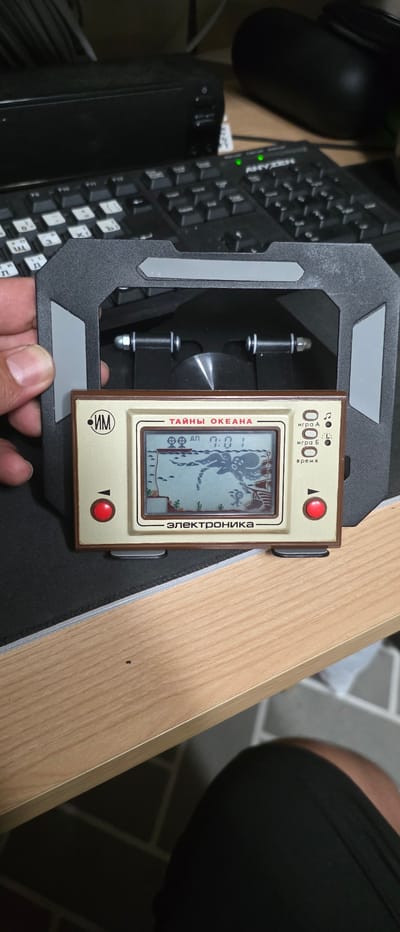 A vintage handheld electronic game, identified by the text "ТАЙНЫ ОКЕАНА" (Mysteries of the Ocean) and "электроника" (Elektronika), is placed on a black mousepad on a wooden desk. The game features a pixelated screen displaying a scene with an octopus and fish, along with the time "0:01". Red buttons are visible on the left and right sides of the game. The game is positioned within a black plastic stand with grey accents. In the background, a black keyboard and a black speaker are partially visible. The image is taken from a slightly elevated angle, and a person's left hand is holding the stand on the left side. The scene is indoors, likely in an office or home environment. There is no visible indication of the city of Gimhae-si, South Korea.