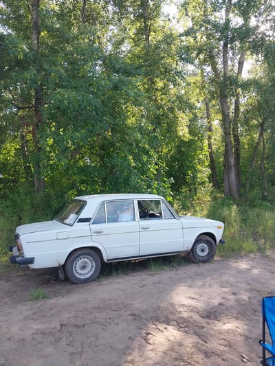 A white, four-door sedan is parked on a dirt path in a wooded area. The car is a classic model, possibly a Lada, with chrome trim and a black bumper. The driver's side window is down, and a person can be seen sitting in the driver's seat wearing a light-colored shirt. Sunlight filters through the dense green foliage of tall trees that surround the scene. The ground is covered in dirt and sparse grass, with some debris scattered about. The overall impression is of a rustic, outdoor setting, possibly a rest stop or recreational area. The image was taken in the city of Chkalov, Russia.