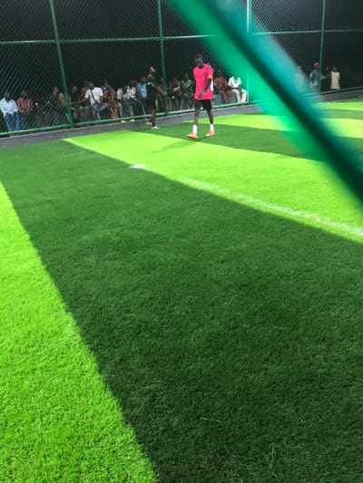 An outdoor soccer field with artificial turf is divided by a white sideline. Several people stand or sit near the fence line on the left. A man in a pink jersey and dark shorts stands on the field, facing the audience. The photo is taken through a green fence, slightly blurring the image. The turf is a vibrant green, with a darker green section.