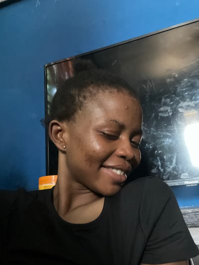 A woman with short, dark hair pulled back in a bun is smiling with her eyes closed. She's wearing a black t-shirt. She appears to be indoors in front of a large TV screen, possibly a Hisense brand, mounted on a blue wall. An orange object is visible near her neck, potentially packaging or a container. The location is Onibudo, Nigeria.