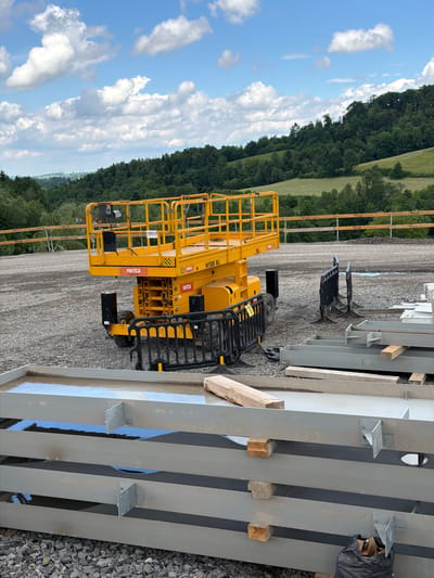 In Wilkowisko, Poland, a yellow scissor lift, identified as H18S XL by Renta, is parked on a gravel surface. The lift is positioned on a construction site, evidenced by stacks of metal beams in the foreground and a temporary wooden fence in the background. The environment is rural, with rolling green hills and a dense forest line under a partly cloudy sky. The scene appears to be a construction project in progress.