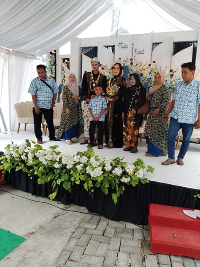 In Juwana, Indonesia, a group of eight individuals, consisting of two children and six adults, is depicted posing for a photograph on a raised white stage. The stage features a black skirt decorated with white flowers and green foliage along its front edge. Behind the subjects, a white backdrop displays the names "Taufiq" and "Eukarti" in blue script, flanked by white and blue floral arrangements. Above the stage, white draped fabric forms a canopy.
At the center, a man and a woman, presumably a bridal couple, are dressed in dark, elaborate traditional Indonesian attire with gold embellishments and headpieces. The woman holds a small bouquet of yellow and white flowers. A young boy in a light blue patterned shirt and dark pants stands directly in front of them.
To the left of the central couple, a man wears a light blue patterned short-sleeved shirt and black pants, carrying a black shoulder bag. Next to him, a woman wears a green, blue, and black patterned long dress and a light beige hijab.
To the right of the central couple, three adults are present. A woman in a dark brown, gold, and black patterned long dress and a dark hijab carries a brown shoulder bag. Beside her, another woman wears a blue, gold, and black patterned long dress and a light brown hijab. On the far right, a man wears a light blue patterned short-sleeved shirt and blue jeans.
On the stage, behind the left-most man, a white decorative chair is visible. The ground level in front of the stage is a mix of concrete and pavers. A green mat is partially visible on the ground to the lower left, and a red fabric-covered two-step platform with a small dark object and a piece of white paper rests on the ground to the lower right.