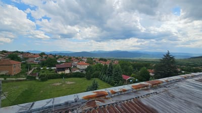 The photograph depicts a view of Hristo Danovo, Bulgaria, from a slightly elevated perspective, possibly from a rooftop. The foreground features a corrugated metal roof in a state of disrepair, exhibiting signs of rust and weathering. Beyond the roof, the scene opens up to a landscape of houses with red-tiled roofs interspersed among green trees and grassy areas, indicating a village or small town setting.
In the middle ground, the village extends toward a broad valley, with progressively fainter details suggesting distance. In the background, a range of mountains is visible under a sky filled with puffy white and grey clouds. The lighting suggests it's daytime, with soft shadows cast by the clouds. There are no people visible in the image. The overall environment is a mix of residential and natural, with the mountains providing a scenic backdrop to the village.