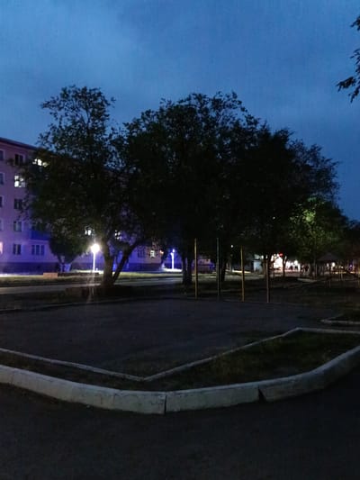 The image depicts an outdoor scene at dusk or night in the city of Gvardeyskiy, Kazakhstan. A multi-story apartment building with illuminated windows is visible on the left side of the frame. In front of the building, a street or pathway is lined with mature trees, some of which are lit by streetlights. The foreground shows a paved area with a raised, concrete border surrounding a patch of grass or soil. In the background, beyond the trees, further buildings and lighting are present, suggesting a residential or mixed-use area. The overall atmosphere is tranquil and somewhat subdued due to the low light conditions.