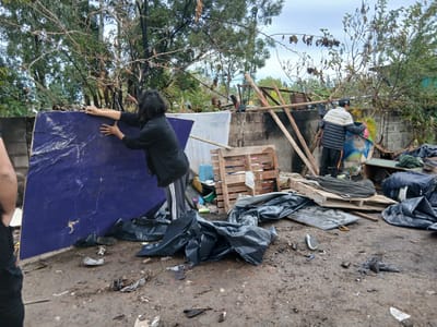 The scene appears to be an abandoned or neglected area in San Fernando, Argentina. The ground is muddy and littered with debris, including pieces of wood, black plastic bags (likely containing trash), and various other discarded items. Two individuals are present. One is holding a large, dark blue sheet or board, while the other is standing near a concrete wall and what appears to be more debris and discarded items. The general atmosphere suggests a state of disrepair and neglect. A concrete wall is visible in the background, partially obscured by debris and vegetation. Trees and a cloudy sky are visible above the wall.