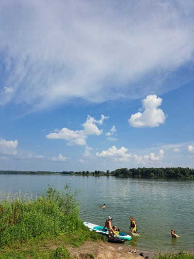 In Sorochii Log, Russia, on a clear day with scattered clouds, a group of people are enjoying the water at a lake. The lake is calm and the shoreline is lined with lush green grass and trees in the distance. A paddleboard is partially submerged in the shallow water near the shore, with a man and two children on or around it. A young boy is reaching for an inflatable tube, while a girl in a life vest is stepping into the water. Another child is swimming in the lake further out, and a person is visible in the water to the right.