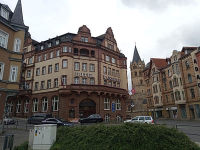 The image captures a street scene in Eisenach, Germany, with a prominent hotel named "HOTEL KAISERHOF" as the central focus. The hotel is a grand, multi-story building with ornate architecture, featuring a light-colored brick facade and dark red stone accents. It has a steeply pitched dark roof with dormer windows. Decorative balconies with flowering plants adorn the upper levels.

Adjacent to the hotel, to the right, is a church tower with a pointed spire and a clock face. The church is constructed of light brown stone. Further to the right, several other buildings line the street, displaying a similar architectural style with varying colors and window designs. Some of these buildings appear to be shops, with visible signage.

In the foreground, a street runs across the scene, bordered by a low black metal fence and a dense green hedge. Several cars are parked and in motion on the street. A dark grey van is parked on the left side of the street, while a dark blue sedan is in motion in the center. A silver station wagon is also visible, driving away from the camera on the right side of the street.

A white electrical box, adorned with graffiti, is situated in the lower left portion of the image, partially obscured by the hedge. The sky is overcast, suggesting a cloudy day. The overall impression is of a historic urban environment.