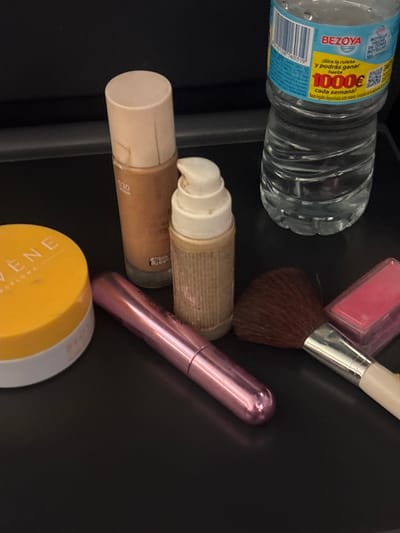 In Santa Maria de Palautordera, Spain, on a dark, flat surface, a collection of cosmetic items and a water bottle are arranged. From left to right, there is a yellow, round container with a white lid and faint text. Next to it, a tall, beige bottle with a beige cap and gold text. To its right, a smaller, textured beige bottle with a white pump dispenser. Further right, a pink metallic tube lies horizontally. To the right of the pink tube, a makeup brush with a dark brown bristled head and a silver ferrule with a white handle is positioned diagonally. Adjacent to the brush is a small, pink rectangular item, possibly blush. In the background, a partially visible clear plastic water bottle with a blue and yellow label is standing upright. The label on the bottle includes text in Spanish. The lighting suggests an indoor setting with diffused light. There are no people visible in the image.