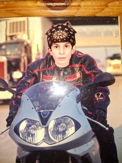 The image is a photograph, framed in wood, showing a young boy wearing a black and red motorcycle jacket and a black bandana. He is sitting on a silver motorcycle, with the front of the motorcycle and his upper body visible. The background is blurred and appears to be an outdoor setting, possibly in a city with buildings and vehicles. There are no discernible elements that specifically identify the location as Medvezhyi Ozyora, Russia. The image is slightly dusty and scratched, suggesting it may be an older photograph.