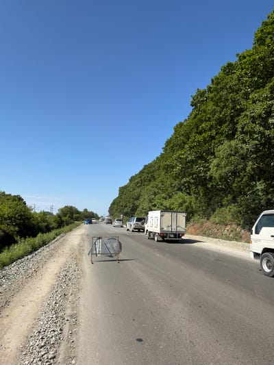 This is a longitudinal, eye-level, medium shot of a road in Vladimiro-Alexandrovskoye, Russia, on a clear day. The road is asphalt, with a gravel shoulder on the left. Lush green trees line the right side of the road, and a dense forest is visible on the hillside. In the foreground, a temporary metal barrier with two reflective signs is positioned on the road. Several vehicles are present, including a white box truck, a white minivan, a silver SUV, and other cars further down the road. Traffic appears to be moving slowly. The sky is a bright, cloudless blue.