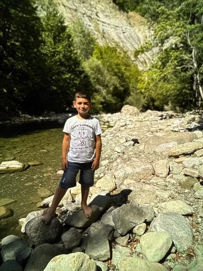 A young boy stands on a rocky riverbank in a wooded area. He is wearing a white t-shirt with a graphic and blue denim shorts. The river is to his left, and a rocky, mountainous incline covered in trees is behind him. The lighting suggests it is daytime, with sunlight dappling through the leaves. There is no specific indication of the city of Artik, Armenia in the image.