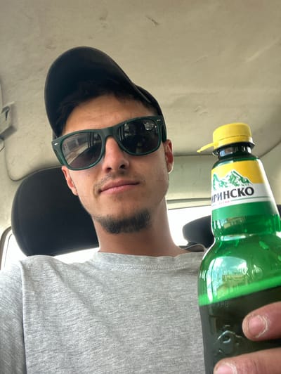 A person is sitting in a car, wearing sunglasses and a black baseball cap. They are holding a bottle of a drink labeled "ПИРИНСКО" (PIRINSKO). The interior of the car is visible with beige-colored panels and a black car seat. The specific location in the city of Isperihovo, Bulgaria is not discernible from the image.
