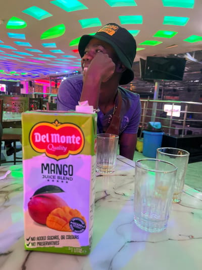 The image, taken in Kimorori, Kenya, depicts a person seated at a table in an indoor setting, likely a lounge or restaurant. The main subject is a dark-skinned male wearing a black bucket hat with partially visible yellow text ("DOG") and a blue or grey t-shirt with a brown shoulder strap. He is looking towards the camera with a slight smile, his right hand positioned near his face.
On the white marbled table in front of him are three empty glasses of different sizes: two larger, clear fluted glasses and one smaller glass. Also prominently featured on the table is a Del Monte Mango Juice Blend carton. Visible text on the carton includes "Del Monte Quality", "MANGO JUICE BLEND", "SWEETENED", "PINEAPPLE", "NATUR", "NO ADDED SUGARS, OR COLOURS", "NO PRESERVATIVES", and "1 LITRE".
The background features a wall adorned with numerous illuminated rectangular segments in green and blue hues, creating a vibrant and decorative light pattern. Other background elements are dimly lit, suggesting an evening or nighttime setting. The scene conveys a casual and relaxed atmosphere, with the person seemingly enjoying a drink in a colorfully lit space.