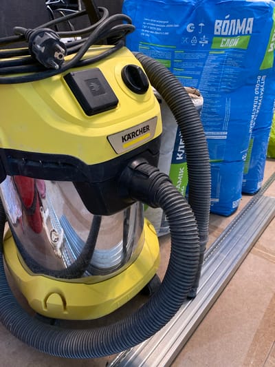 A Karcher wet/dry vacuum cleaner, yellow and chrome, sits on a light brown surface. A black power cord is coiled on top of the vacuum. To the right of the vacuum are two large blue bags of building material. A metal track lies horizontally in the foreground. The background is dark and out of focus. There are no people in the image. The scene appears to be indoors, possibly a workshop or construction area. The location is a dwelling in the village of Peschanie Kovali, Tatarstan, Russia.
