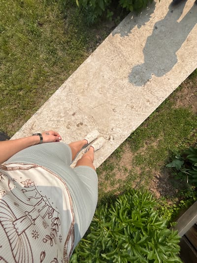 A first-person perspective shot shows a person's legs and torso standing on a concrete path outdoors, with a grassy area to the left and the right. The person is wearing light-colored sneakers and a pale blue ribbed outfit with a brown graphic print on the front. A watch is visible on the person's left wrist, and a small sun tattoo is on the back of their hand. A shadow of the person is cast on the concrete path. Lush green plants and grass are present in the surrounding environment. The location is identified as Tartat, Russia.