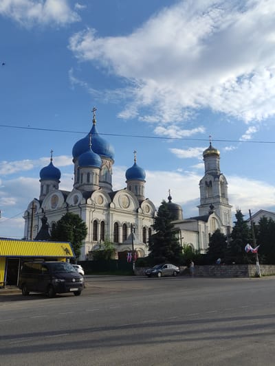 The image shows a large white church with multiple blue domes and a single gold dome on a bell tower, located in the city of Rogachovo, Russia. The church architecture features intricate detailing, arched windows, and crosses on its domes. In the foreground, a black van is parked in front of a yellow building with a corrugated metal roof. Across the street, a silver sedan is parked and a person is standing near a low brick wall decorated with a wreath. Several evergreen trees are scattered around the church and the surrounding area. The sky is blue with scattered white clouds, and a few birds are visible in the sky. Power lines stretch across the upper portion of the image. The road in front of the church and the yellow building appears to be asphalt. The scene depicts an exterior view of a religious site and its immediate surroundings on a clear day.