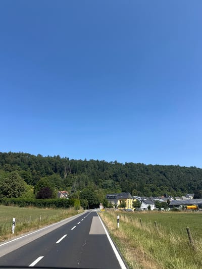 The image shows a road winding through a rural landscape in Schoenfels, Luxembourg. The road is paved with asphalt and marked with white dashed lines. On either side of the road are grassy fields and trees. In the distance, there are houses and buildings, some with solar panels on their roofs. A dense, green forest covers the hillside in the background. The sky is a clear, bright blue. The scene appears to be a sunny day.