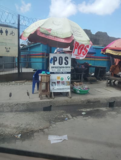 The image depicts a street scene in Oworonshoki, Nigeria, under a cloudy sky. In the foreground, a concrete sidewalk and gutter are visible, with some debris scattered on the ground.
The main subject is a roadside stall or booth. A large, open umbrella, predominantly white and red, is set up to provide shade. A sign hanging from the umbrella reads "POS IS AVAILABLE HERE" in large red letters. Below this, another sign indicates "SIM REGISTRATION CENTER, WIFI AND ROUTER AVAILABLE HERE". This sign also features logos for mobile network providers like GLO and Airtel, along with images related to prepaid and postpaid services.
Adjacent to the POS stall, a simple wooden stand supports a small object, possibly a cash register or payment terminal. A blue plastic chair is placed nearby.
In the background, a chain-link fence with barbed wire runs across the scene. Behind the fence, a blue building with colorful horizontal stripes is partially visible. Further back, the landscape appears somewhat rugged with hints of other structures. Another umbrella, partially visible on the right side of the image, is a vibrant pink, suggesting another vendor or stall.
The scene suggests a typical urban commercial environment, likely a busy area where people can access mobile services and make electronic transactions. There are no people clearly visible in the frame.