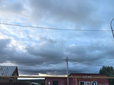 This is a wide shot of a cloudy sky over a rural landscape in Valy, Russia. The sky is filled with a mix of dark, ominous storm clouds and patches of lighter blue sky, suggesting an approaching storm. A power line stretches horizontally across the frame, bisecting the sky.
In the foreground, there are two buildings. On the left, a small building with a rusty, corrugated metal roof is partially visible. In the center and right, a larger, single-story building with a reddish-brown facade and a dark roof is prominent. This building appears to be a shop or cafe, as indicated by the visible text "КАФЕ" (KAFE, meaning Cafe in Russian) written in white block letters above two windows. The windows themselves have visible signs and some reflections. A yellow pipe or railing runs horizontally in front of this building.
A utility pole with wires is situated in front of the reddish-brown building, adding to the sense of a built-up, possibly somewhat utilitarian, environment. In the background, a dense line of trees and a few distant structures can be seen, fading into the overcast horizon. The overall mood is somewhat somber and dramatic, due to the heavy clouds. The time of day appears to be late afternoon or early evening, given the quality of light.