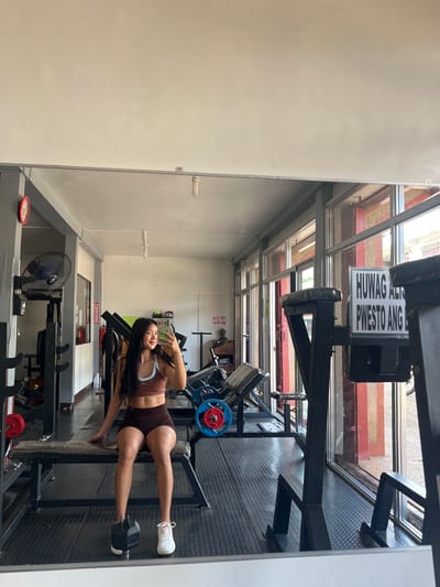 The image depicts a young woman taking a mirror selfie in a gym. She is wearing a brown sports bra and matching shorts, with white sneakers. She is seated on a weight bench in the foreground, with a black dumbbell placed on the floor next to her left foot. The gym appears to be indoors, with gym equipment visible in the background, including weight machines and a stationary bike. There are large windows on the right side of the image, revealing a street scene outside. A sign with Filipino text is visible on a piece of gym equipment. The overall lighting suggests it is daytime. The location is identified as Cuyo, Philippines.