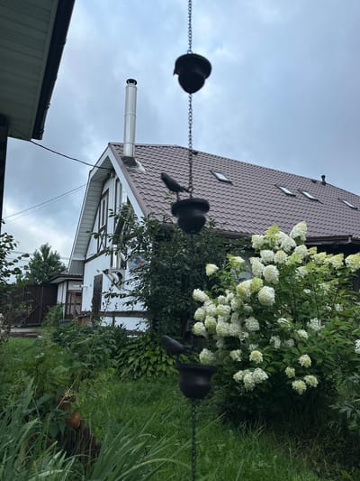 The image depicts a backyard scene in Ромашкино, Russia. A two-story house with a white facade, timber framing accents, and a brown tiled roof dominates the background. A silver chimney pipe extends from the roof, and several skylights are visible. In the foreground, a lush garden with green grass and various plants, including a large bush with white flowers, is present. A hanging wind chime with dark elements is in the left portion of the frame. The sky is overcast and gray.