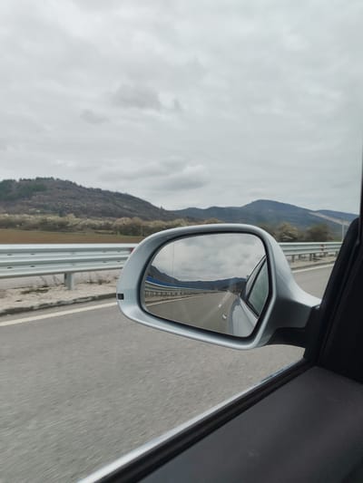 A view from inside a moving car, specifically through the driver's side window showing the side mirror. The reflection in the side mirror shows a highway with a grey sky above. Outside of the car, the image includes a guardrail, a highway, and distant mountains under a grey, cloudy sky. The location is in the region of Novachene, Bulgaria, as indicated by the landscape.