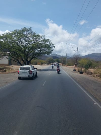 The image captures a road scene in Guatacaral, Venezuela, featuring a white Hyundai Tucson driving on the left side of the road. Ahead of the car, a group of motorcycles are moving in the same direction. The road is bordered by gravel on the right and a large tree and a building on the left. Power lines stretch across the sky above the road. In the background, mountains are visible under a sky with scattered clouds.