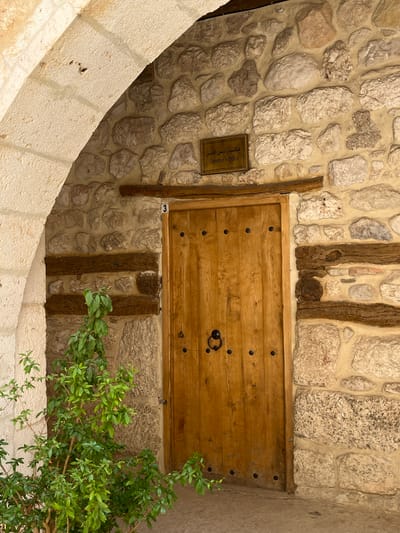 The image depicts a wall and arched walkway in Ma'loula, Syria. The wall is constructed of irregularly shaped stones with wooden beams. A wooden door with metal studs is set into the wall, and a small sign reading "Superior's Office" is visible. A green plant grows near the door. The walkway is made of light-colored stone.