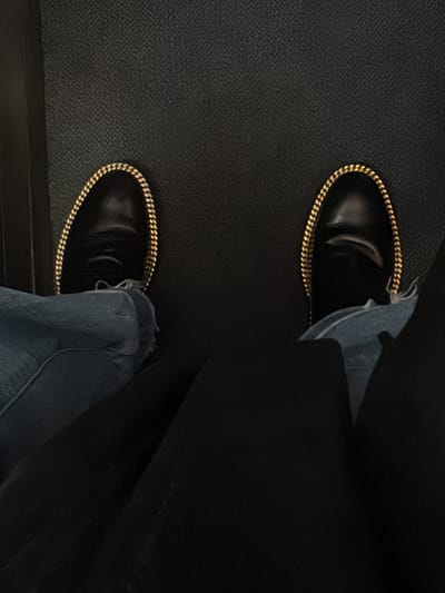 The image presents a top-down view of a person's feet and lower legs. The individual is wearing black, possibly leather, slip-on shoes adorned with a prominent gold chain detail that outlines the top opening of each shoe. The gold chains consist of numerous small, linked rings, catching the light brightly against the dark shoe material.

Above the shoes, the cuffs of light blue denim jeans are visible on both legs. The jeans appear to have a slightly distressed or frayed edge, particularly noticeable on the right foot's cuff. The denim fabric is creased, suggesting the person is in a seated position. The upper portion of the legs, beyond the denim jeans, is covered by a dark, solid-colored fabric, which appears to be part of trousers, a skirt, or perhaps a blanket, creating a contrast with the lighter blue denim.

The setting appears to be indoors, with a dark, textured floor (possibly carpet or a mat) serving as the background. The texture of the floor is a fine, repeating pattern, almost like a cross-hatch or woven design. The lighting is somewhat dim but sufficient to highlight the metallic gleam of the gold chains on the shoes. The overall impression is of a casual yet stylish outfit, viewed from the wearer's perspective in a relaxed, stationary posture. No text is visible in the image.