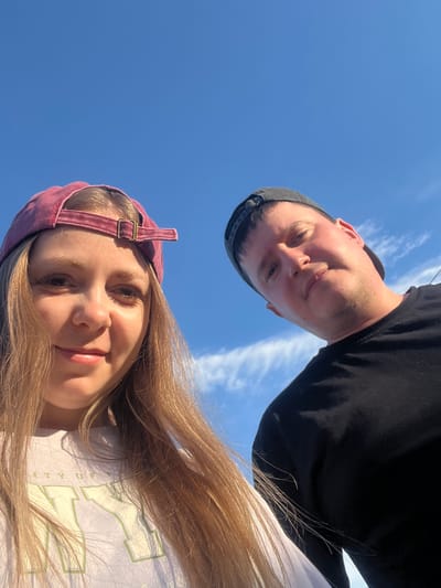 A low-angle, close-up selfie features two individuals against a bright blue sky with scattered white clouds. On the left is a woman with long, blonde hair, wearing a maroon baseball cap and a light-colored t-shirt with green lettering. She is looking towards the camera with a slight smile. To her right and slightly behind is a man wearing a dark baseball cap and a black t-shirt. He is also looking towards the camera, his expression neutral. The camera appears to be held from below, creating an upward perspective of the subjects and the sky. The image is taken outdoors, likely in the city of Mal'tsevo, Russia, given the context.