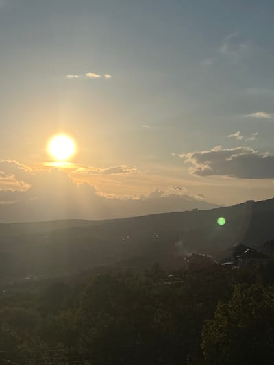 The image shows a landscape during sunset or sunrise. The sun is positioned in the lower left quadrant, emitting a bright glow. Clouds are partially illuminated by the sun's light, forming an arc towards the upper center of the image. A mountain ridge occupies the right side, with some indistinct buildings and possibly dense vegetation towards its base. The overall environment appears hazy or misty.

Without more contextual information, such as clear landmarks or features unique to Kribul, Bulgaria, it is difficult to verify the location within that city.