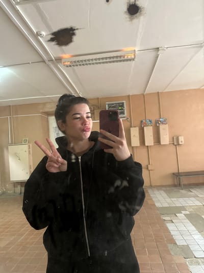 A young woman with dark hair pulled up in a bun is taking a mirror selfie. She is wearing a black zip-up hoodie and is making a kissing face while holding up a peace sign with her left hand. She is holding a dark purple smartphone in her right hand. The background appears to be an indoor hallway with tiled floors and beige walls. There are several electrical boxes and light fixtures visible on the walls and ceiling. The ceiling shows some water damage and discoloration. A bench is visible against the far wall. The floor tiles are a mix of reddish-brown and white squares, with some areas appearing to be under repair. The scene is likely in a public building or facility.