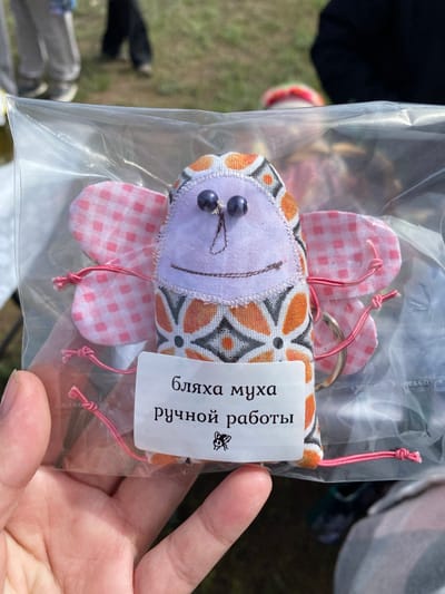 A person's hand holds a sealed plastic bag containing a handmade fabric butterfly keychain. The butterfly has a white face with two dark beads for eyes and a stitched smile, a patterned orange and black body, and pink and white checkered wings. The keychain has a metal ring attached to the top of the butterfly's body. A white tag with text in Cyrillic, including the words "ручной работы" (handmade), is attached to the keychain. The background is blurred, but shows hints of green grass and possibly other people in an outdoor setting. The location is Kumakskoe, Russia.