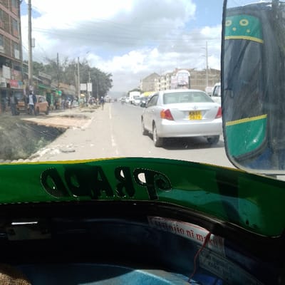 The image presents a forward-facing view from inside a vehicle, revealing a bustling street in Ongata Rongai ward, Kenya. In the immediate foreground, the vehicle's dashboard or interior frame is visible, featuring a green and yellow color scheme with the word "PRADO" in reverse script. A green and yellow side mirror with star decals partially obstructs the view on the far right.

A two-lane paved road, marked with a white center line, extends into the hazy distance under a partly cloudy sky. Directly ahead, a silver Toyota Allion sedan, bearing Kenyan license plate KBS 587Y, occupies the right lane, heading away from the camera. Several other vehicles, including cars and vans, are visible further down the road, indicating moderate traffic flow.

On the left side of the road, a dirt and gravel shoulder contains visible debris. A rudimentary wooden plank bridge spans an uneven section of ground, leading to a pedestrian zone where multiple individuals are visible walking or standing. This area is lined with various commercial structures, including single-story shops with corrugated iron roofs and multi-story buildings, some appearing residential. Utility poles with multiple overhead power lines run along this side of the street.

On the right side of the road, in the mid-distance beyond the silver vehicle, multi-story buildings are observable, one featuring prominent billboards, including an advertisement for "STAREHE." The overall environment suggests an urban or peri-urban commercial and residential thoroughfare.