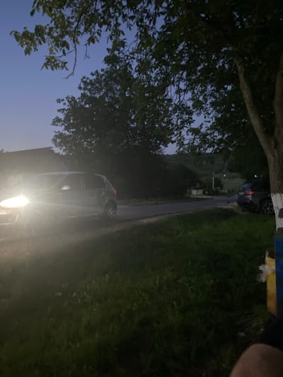 The image shows a street scene at dusk in Negrești, Moldova. A car with its headlights on is parked on the left side of the road, illuminating the foreground with a bright light. In the background, a road curves through a rural landscape with trees and rolling hills. Another car is parked on the right side of the road. The sky is a deep blue, indicating the end of daylight. The overall scene suggests a quiet, rural evening in a Moldovan village.