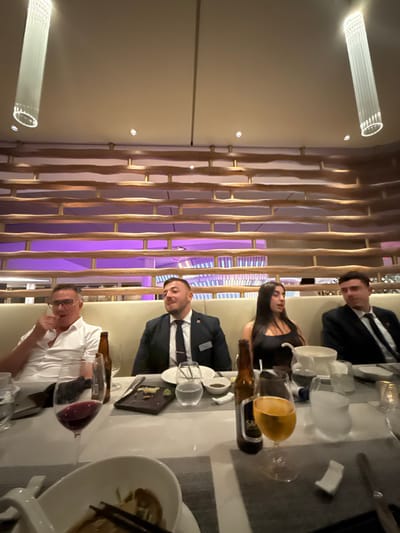 The scene depicts four individuals seated at a dining table within a restaurant or similar establishment in Rum Cay, Bahamas. The environment is illuminated by artificial lighting, including cylindrical pendant lights and recessed spotlights on a light-colored ceiling, suggesting an evening or nighttime setting.
From left to right, four people are visible:
1. A male, wearing a white collared shirt and eyeglasses, is seated with his left arm raised and hand near his chin, appearing to be in conversation or contemplation.
2. A male, wearing a dark suit, white shirt, and dark tie with a tie bar, has a name badge clipped to his lapel. He is looking to his left (viewer's left).
3. A female, with long dark hair, is wearing a dark, possibly sleeveless, top. She is looking to her right (viewer's right).
4. A male, wearing a dark suit, white shirt, and dark tie, is seated at the far right and is also looking to his left (viewer's left).
The table is set with multiple place settings. Visible items include various plates, some containing remnants of food like sushi or small dishes, and chopsticks resting on small holders. Beverages present include a glass of red wine, a glass of amber-colored liquid (likely beer or another alcoholic drink), water glasses, and two dark-colored beer bottles, one partially obscured by the glass. A white bowl with chopsticks and food is prominent in the foreground.
The background features a decorative wall with horizontal, undulating wooden or bamboo-like slats, set against a lighter wall with a distinct purple ambient light emanating from a section further back. The seating appears to be a continuous upholstered booth.