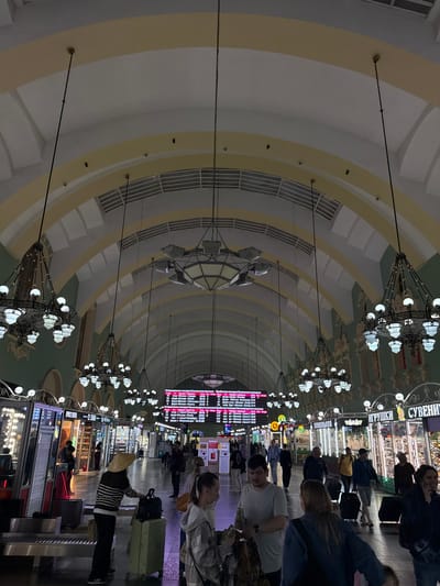 Here is a detailed description of the image:
**General Description:**
The image depicts the interior of a grand railway station concourse, likely a main hall, characterized by its high, arched ceiling, ornate chandeliers, and a bustling atmosphere. The photograph is taken from a standing level, looking lengthwise down the hall, which gives a strong sense of depth and perspective. The orientation of the image is such that the ceiling dominates the upper portion of the frame.
**Environment and Architecture:**
* The ceiling is the most striking feature, featuring a series of arches decorated with pale yellow and white paint and molding. These arches create a tunnel-like effect leading down the hall.
* Large, decorative chandeliers are suspended from the ceiling at regular intervals, providing illumination and adding to the elegance of the space.
* On either side of the hall, there are shops and services catering to travelers.
* Large display boards, likely displaying train schedules and other travel information, are visible at the far end of the concourse.
**People:**
* Many people are present in the hall, either standing or walking. Some are seen with luggage, suggesting they are in transit.
* People appear to be of diverse ages and appearances.
* Some individuals are engaged in conversations, while others seem to be focused on their destinations.
**Objects:**
* Luggage is seen in various forms, including suitcases, backpacks, and bags.
* Signage for shops and services is visible, indicating the presence of businesses within the station.
* The chandeliers themselves are notable objects, contributing to the aesthetic of the space.
* Ticketing booths are visible
**Actions and Interactions:**
* People are primarily engaged in movement, either walking through the hall or pausing to consult information.
* Some individuals are likely purchasing tickets or other travel services.
**Location:**
I am unable to confirm the image's location as Ukromne, Ukraine with certainty, because this would necessitate a more thorough examination of the image's features such as the language on signs or the architectural style.