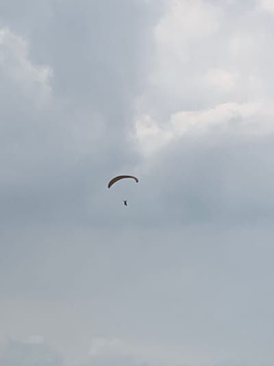 An airborne paraglider is observed against an overcast sky in Puncak, Indonesia. The paraglider features a brown-orange colored canopy in an open, arc-like configuration. Suspended directly beneath the canopy is a single individual, visible as a dark, silhouette-like figure. The sky is predominantly light gray, with patches of darker gray clouds scattered across the frame, indicating an overcast or partly cloudy condition. No other objects, persons, or ground features are visible within the image frame. The individual is actively engaged in paragliding, positioned centrally within the sky.