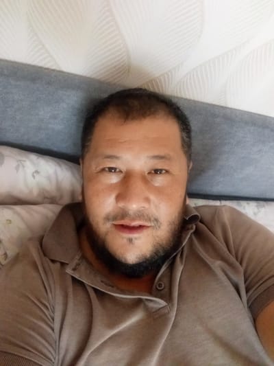 A close-up, top-down selfie shows a man lying on a bed. He has short, dark hair, dark eyes, and a full beard. He is wearing a brown polo shirt with two buttons at the collar. The background consists of a gray headboard and a white textured wall or headboard with a patterned design. There are also pillows visible with a floral pattern in white and light pink. The lighting suggests it is daytime, and the indoor setting implies he is at home or in a hotel. The man's expression is neutral. There is no visible text in the image.